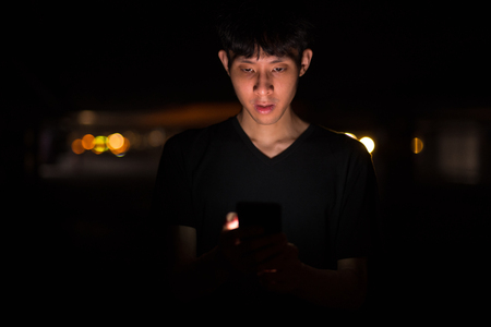 Portrait Of Asian Man Outdoors At Night In Parking Lot Using Mobile Phoneの写真素材