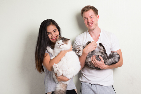 Young happy multi-ethnic couple holding two cats together against white backgroundの写真素材