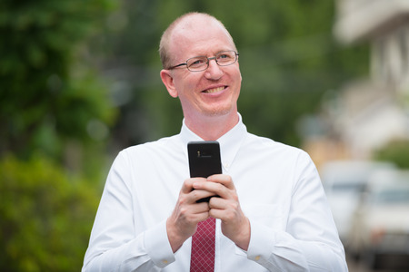Portrait of happy mature bald businessman using phone while thinking outdoorsの写真素材