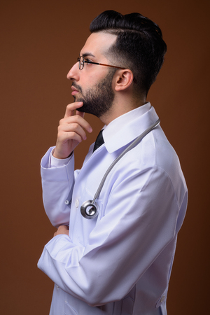Young handsome bearded Iranian man doctor against brown backgrouの写真素材