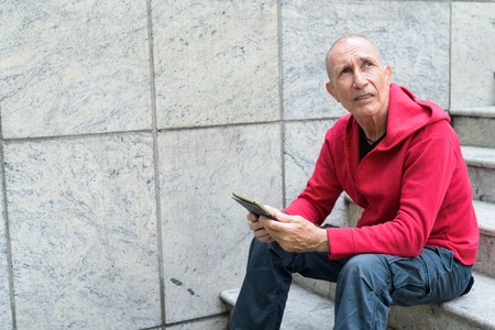 Bald senior man thinking and looking up while holding digital ta