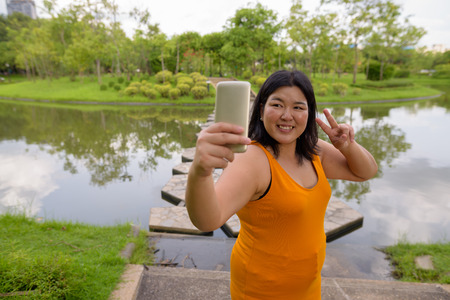 Beautiful overweight Asian woman taking selfie with mobile phone in parkの写真素材