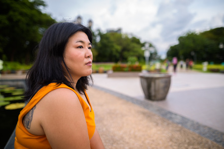 Profile view of overweight Asian woman sitting and thinking in parkの写真素材