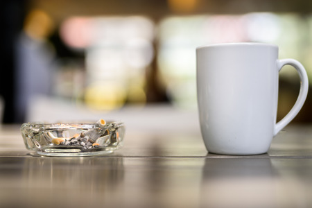 Coffee cup and ashtray with cigarettes on wooden table in restaurant with out of focus background and bokeh effectの写真素材