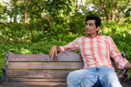 Indian man thinking while sitting on wooden bench in peaceful grの写真素材