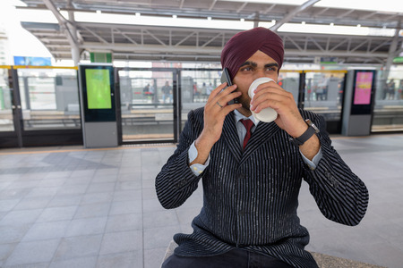 Indian businessman talking on phone at train station while drinking coffeeの写真素材