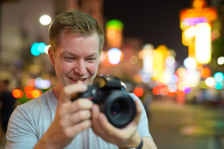Face of young happy tourist man photographing with camera in the streets of Chinatown at nightの写真素材