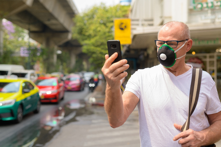 Senior man using face mask to protect from pollution smog in city while holding mobile phoneの写真素材