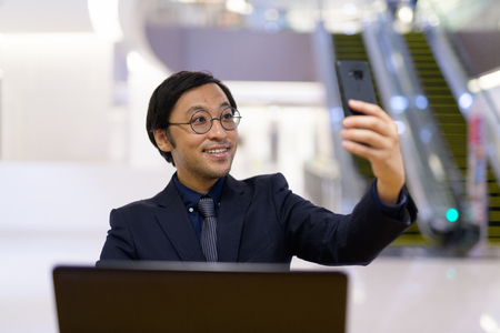 Happy Asian businessman using laptop and taking selfie inside the office buildingの写真素材
