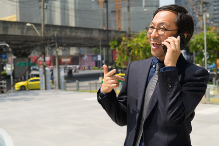 Happy Asian businessman talking on the phone in the city streets outdoorsの写真素材