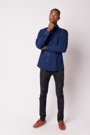 Full body shot of young handsome bald African businessman thinkingの写真素材