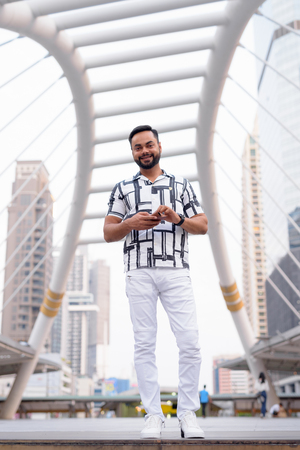 Full body shot of happy young bearded Indian man smiling in the cityの写真素材