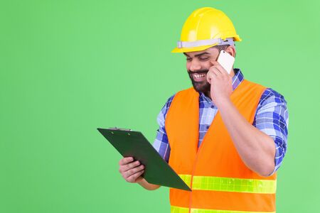 Happy young overweight bearded Indian man construction worker reading on clipboard and talking on...