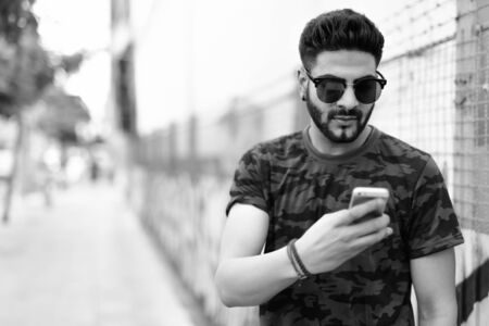 Young handsome Indian man wearing sunglasses while using mobile phone against dirty urban wall with...