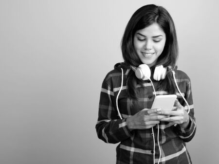 Young beautiful Indian woman wearing headphones and using mobile phone against gray backgroundの写真素材