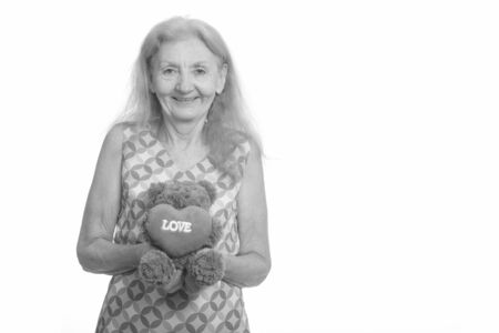 Studio shot of happy senior woman smiling while holding teddy bear with heart and love signの写真素材