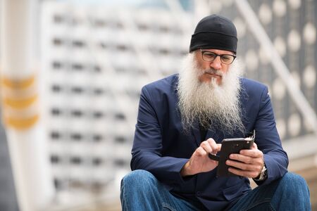 Mature bearded businessman sitting while using phone in the city outdoors