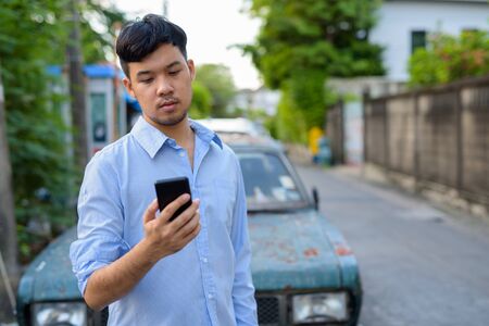 Young Asian businessman using mobile phone against rusty old car