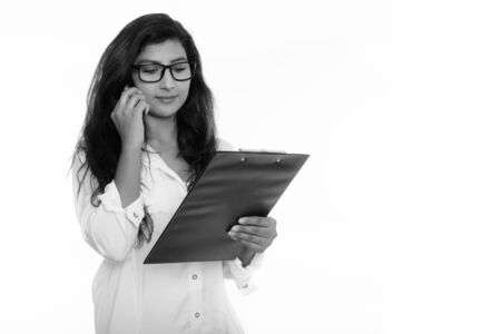 Studio shot of young beautiful Persian woman talking on mobile phone while reading on clipboardの写真素材