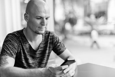 Young handsome bald man using mobile phone inside restaurant with glass windowsの写真素材