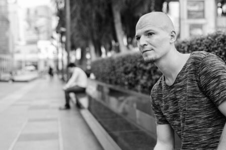 Young handsome bald man thinking while sitting on concrete bench outside of the mallの写真素材