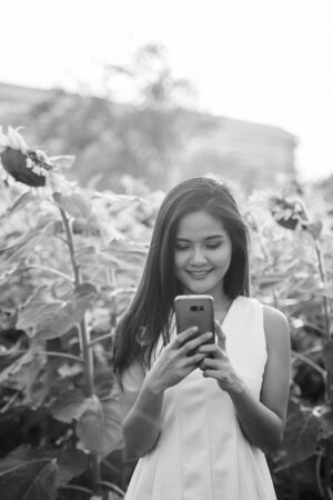 Young happy Asian woman smiling while using mobile phone in the field of blooming sunflowersの写真素材