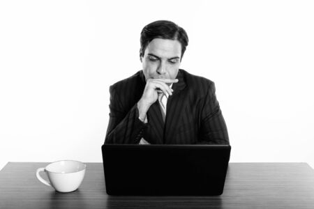 Studio shot of young Persian businessman thinking while using laptop with coffee cup on wooden table...