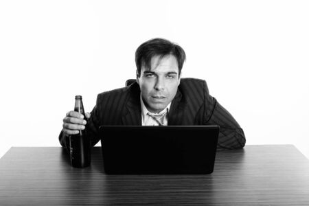 Studio shot of Persian businessman looking drunk while holding bottle of beer and looking at camera with laptop on wooden tableの写真素材