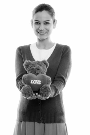 Studio shot of happy young beautiful woman smiling while holding teddy bear with heart and love signの写真素材