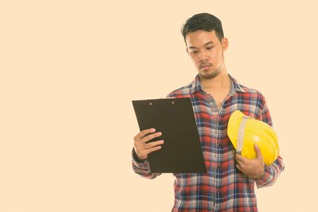 Studio shot of young Asian man construction worker reading on clipboard with holding safety helmet
