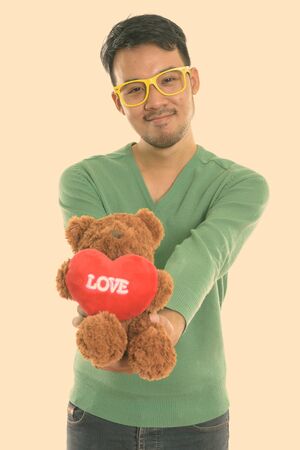 Studio shot of young Asian man giving teddy bear with heart and love signの写真素材