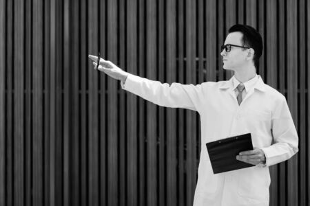 Profile view of young handsome man doctor holding clipboard and pen while pointing finger outdoorsの写真素材