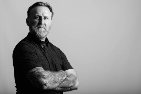 Studio shot of mature bearded man with hand tattoos with arms crossed in gray backgroundの写真素材