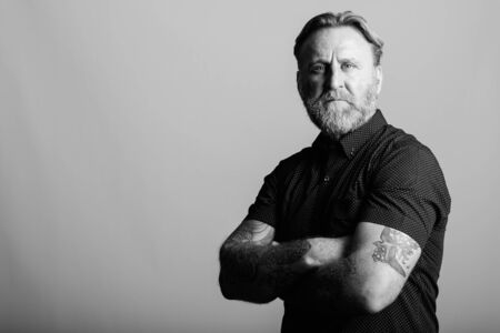 Studio shot of mature bearded man with hand tattoos with arms crossed in gray backgroundの写真素材