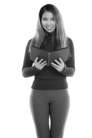Studio shot of young happy Asian woman smiling and standing while holding bookの写真素材