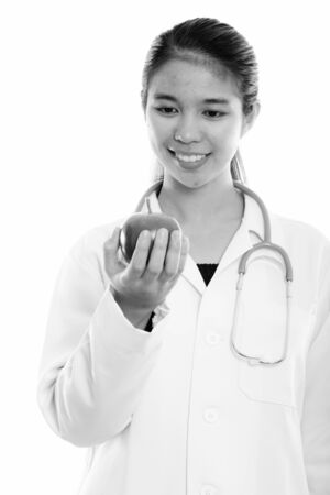 Studio shot of young happy Asian woman doctor smiling while holding and looking at red appleの写真素材