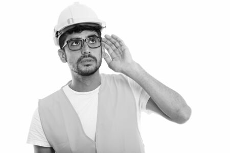 Studio shot of young Persian man construction worker listening while thinking and looking upの写真素材