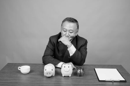 Mature Asian businessman sitting with wooden table against gray backgroundの写真素材