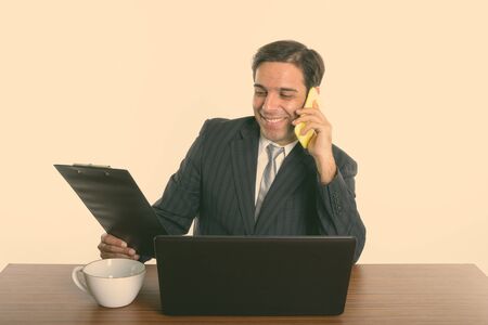 Happy Persian businessman talking on the phone while reading on clipboard on wooden table