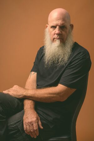 Mature bald man with long gray beard against brown background