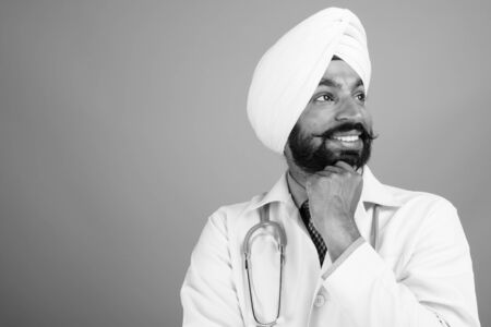 Studio shot of young handsome bearded Indian Sikh man doctor against gray background in black and...