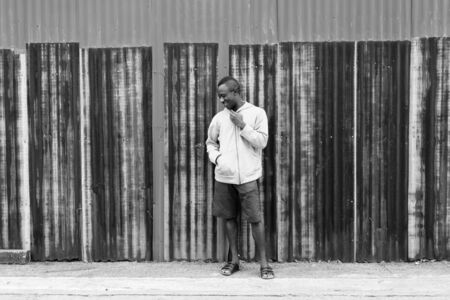 Young happy black African man smiling and thinking while holding jacket against old rusty sheet wallの写真素材