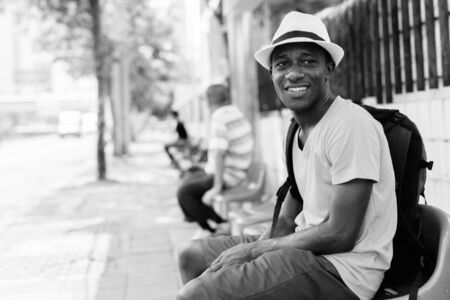 Happy young African tourist man smiling and thinking while sitting at the bus stop