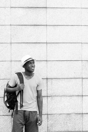 Happy young African tourist man thinking and holding backpack against concrete block wallの写真素材