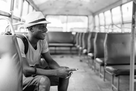 Profile view of young happy African tourist man using mobile phone while riding the busの写真素材