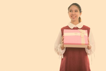 Studio shot of young happy Asian woman smiling while holding gift box and thinkingの写真素材