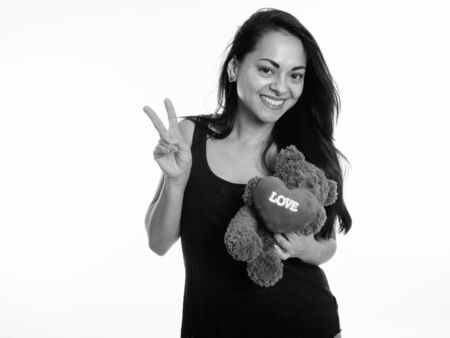 Studio shot of happy beautiful woman smiling and giving peace sign while holding teddy bear with heart and love sign against white backgroundの写真素材