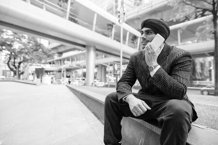 Young handsome Indian Sikh businessman talking on the phone in the city outdoors