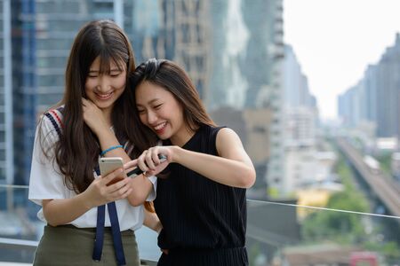 Two happy young beautiful Asian teenage girls using phone together against view of the cityの写真素材