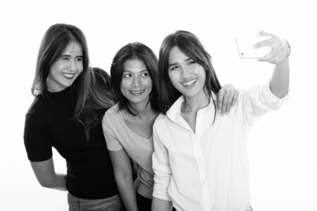 Studio shot of three happy young Asian woman friends smiling while taking selfie picture together with one mobile phoneの写真素材
